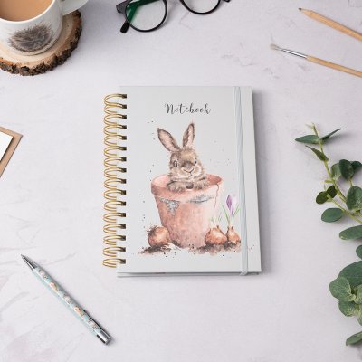 A small Rabbit inside a flower pot peaking over the edge on an A5 Spiral Bound Notebook surrounded by stationary and a hot drink