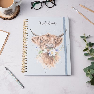 A highland cow chewing a mouthful of flowers on an A4 spiral bound notebook on a desk surrounded by stationery