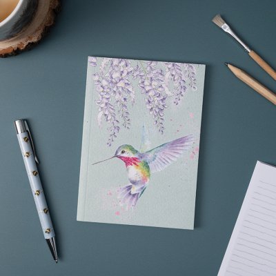 A colourful hummingbird flying on an A6 paperback notebook on a desk surrounded by stationery.