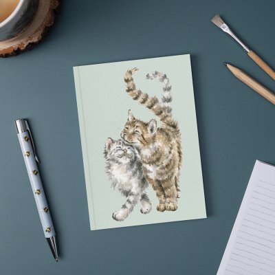 Cuddling cats illustration on A6 paperback notebook on desk surrounded by a pen and pencil 
