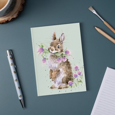 Rabbit eating clovers illustration on A6 paperback notebook on a desk surrounded by pen and pencil 
